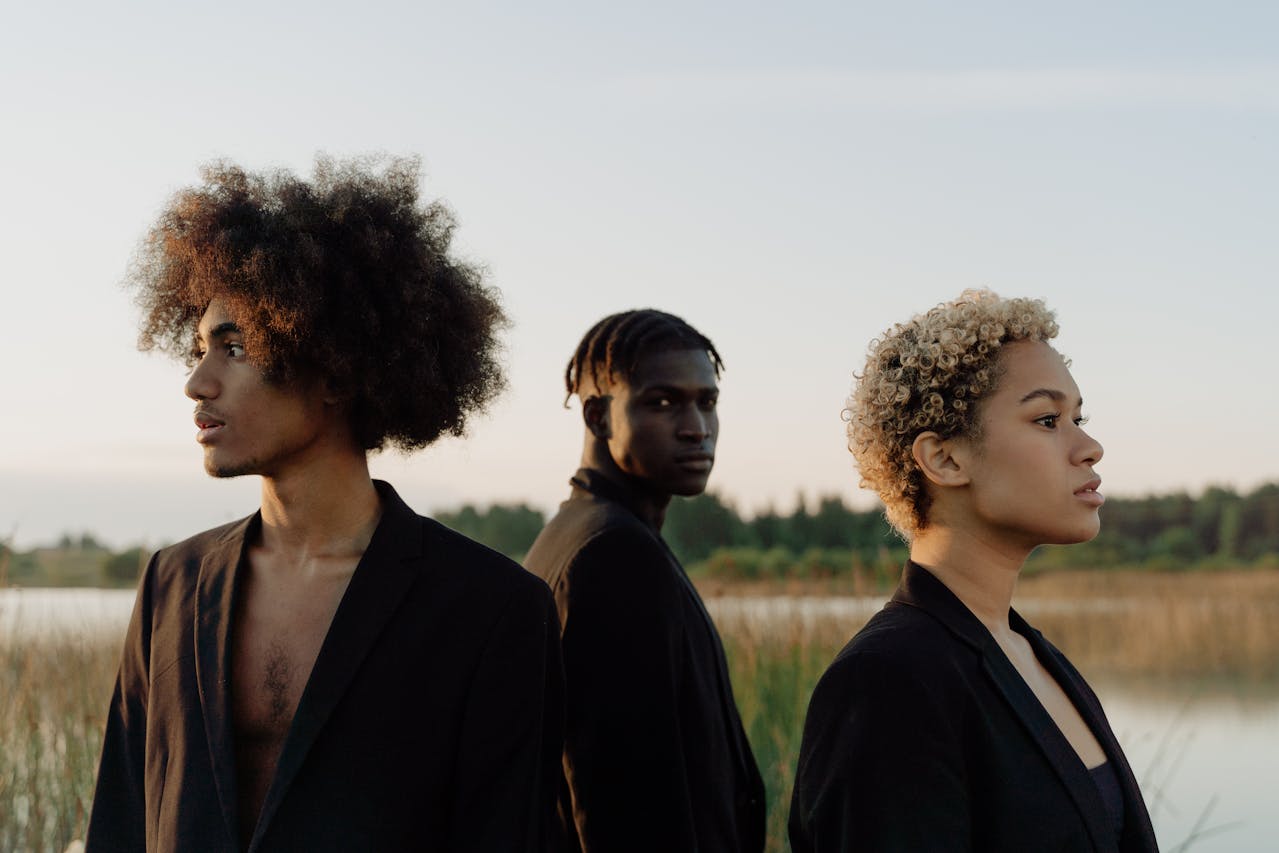 Drie stijlvolle jonge mensen met diverse haartypes in zwarte outfits, staand in een natuurlijk landschap bij het water tijdens zonsondergang, kijkend in verschillende richtingen met een serene en krachtige uitstraling.